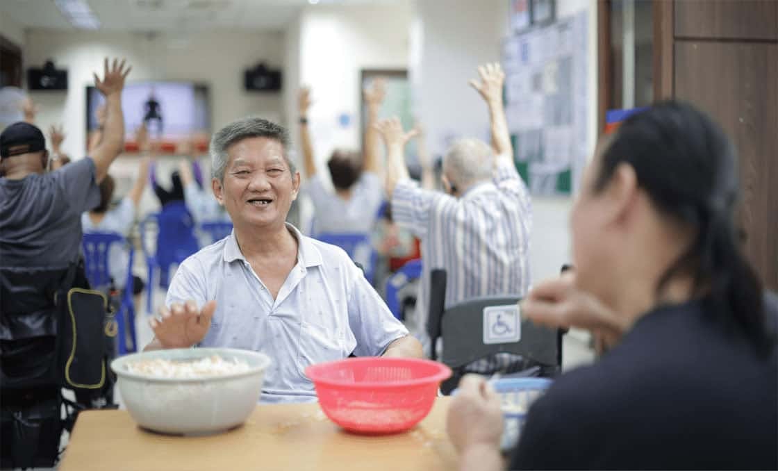 An elderly man prepping bean sprouts in a community centre, behind him, a group of elderlies are seated and exercising with their arms in the air
