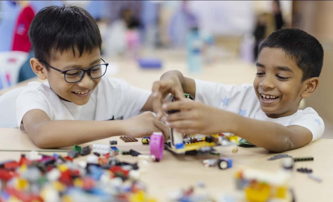 Two smiling boys playing and building an unidentified object with lego blocks